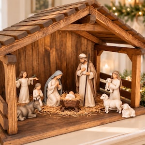 May include: A wooden nativity set featuring a stable structure housing figurines of Mary, Joseph, the infant Jesus in a manger, an angel, a child, a donkey, and two sheep. The figures are rendered in neutral tones with soft fabric-like textures.
