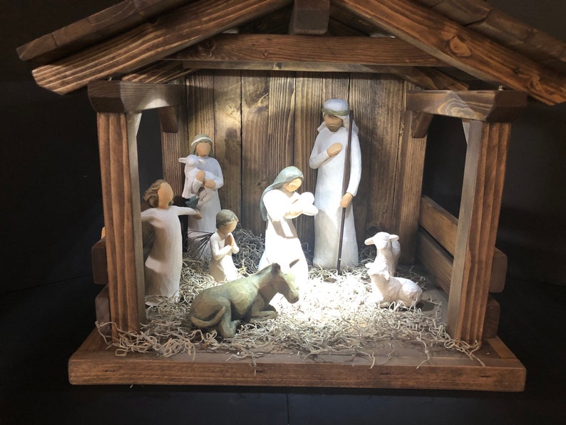 May include: A wooden nativity scene with figures of Mary, Joseph, baby Jesus, and other figures. The figures are in white robes, and the scene is set in a wooden stable with a hay-like base. The overall color scheme is neutral with warm lighting.