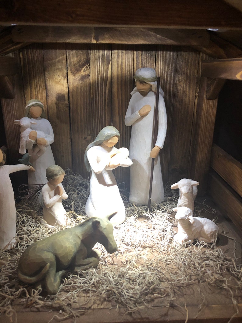 May include: A wooden nativity scene featuring carved figures of Mary, Joseph, baby Jesus, shepherds, angels, and animals. The figures are in white and beige tones, set inside a rustic wooden stable with straw.