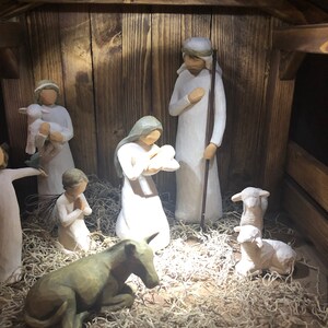 May include: A wooden nativity scene featuring carved figures of Mary, Joseph, baby Jesus, angels, shepherds, and animals. The figures are in white and beige tones, set inside a rustic wooden stable with straw bedding.