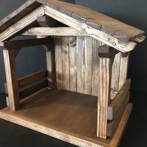 May include: A rustic wooden nativity stable with a pitched roof. The structure is made of stained wood, featuring a base, walls, and a roof. The design includes a small interior space, with visible wood grain and construction details.