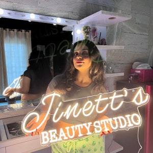 May include: A woman holds a white neon sign that reads "Jimett's Beauty Studio". The sign is shaped like a state of Georgia. The woman is wearing a green and white patterned shirt.