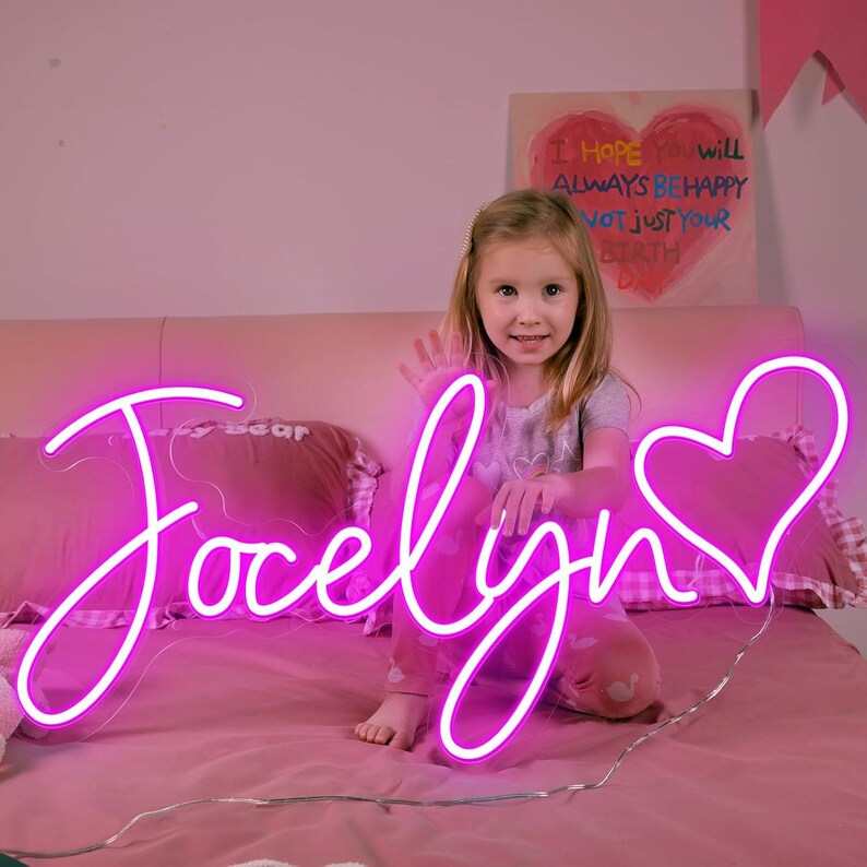 May include: A bright pink neon sign spelling "Jocelyn" with a heart symbol. The sign is on a pink bed with a young girl in the background. A painting with the words "I HOPE YOU WILL ALWAYS BE HAPPY NOT JUST YOUR BIRTH" is visible.