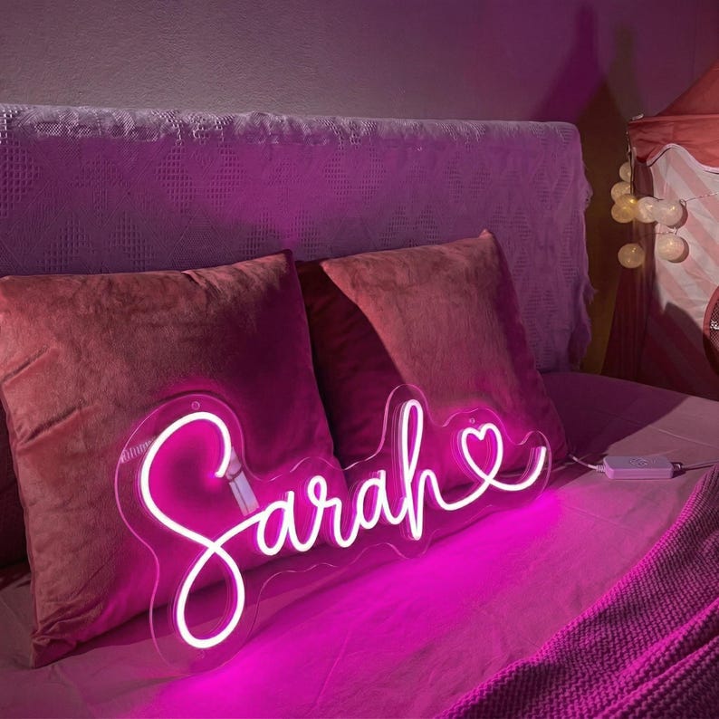 May include: A neon sign in the shape of the name "Sarah" in a cursive font, with a heart symbol at the end. The sign emits a bright pink glow and is placed on a bed with pink pillows and bedding.