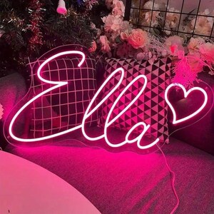 May include: A neon sign in a cursive font spelling "Ella" in bright pink. A heart shape is at the end of the name. The sign is on a dark gray couch with patterned pillows and flowers in the background.