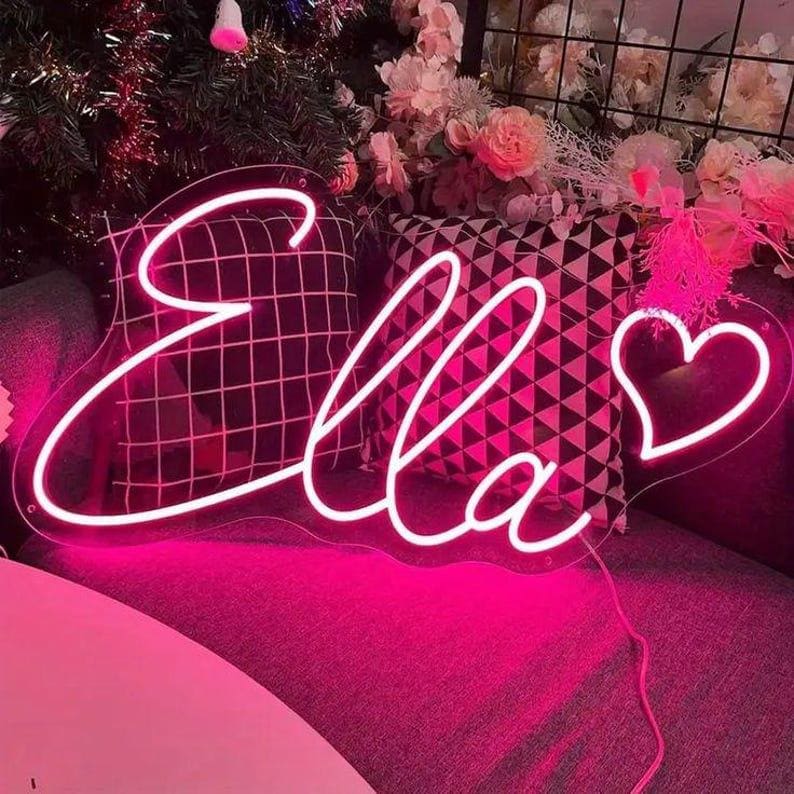 May include: A neon sign in a cursive font spelling "Ella" in bright pink. A heart shape is at the end of the name. The sign is on a dark gray couch with patterned pillows and flowers in the background.
