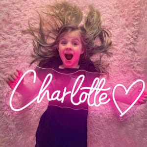May include: A neon sign in the shape of the name "Charlotte" with a heart symbol. The sign is lit with a bright pink light. The sign is held by a person lying on a pink fluffy surface.