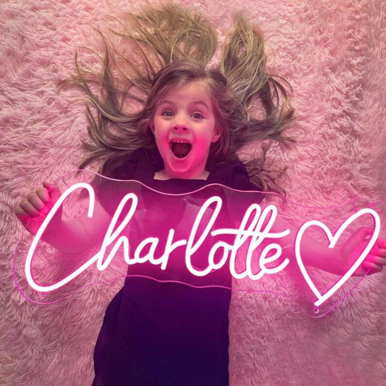 May include: A neon sign in the shape of the name "Charlotte" with a heart symbol. The sign is lit with a bright pink light. The sign is held by a person lying on a pink fluffy surface.