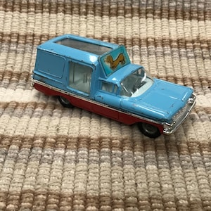 May include: A vintage toy delivery truck in light blue and red. The truck has a clear roof and a sign with a dachshund on top. The toy is on a striped beige and brown surface.