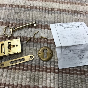 May include: A collection of brass hardware components, including a key, lock mechanism, escutcheon, and screws. A paper with the text "DOOR OR DRAWER LOCK" is also present. The hardware is likely for a cabinet or drawer.