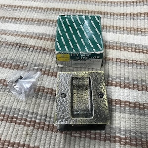 May include: A bronze-toned, rectangular door pull with a textured surface, a black base, and a matching box labeled "SLIDE-LOK". A small bag of screws is also present.