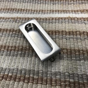 May include: A rectangular, brushed-metal recessed pull with a curved interior. The hardware is designed to be flush with a surface. The pull is silver and has a small screw on the bottom.