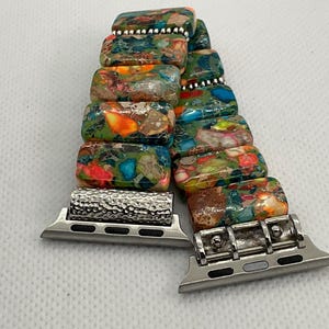 May include: A vibrant watch strap featuring rectangular, multi-coloured stone beads in shades of green, orange, red, and brown. The strap has silver-coloured metal connectors.