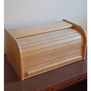 May include: A light-colored wooden bread box featuring a horizontal roll-top door mechanism. The rectangular storage container has smooth side panels and a slatted front design that slides upward to reveal the interior compartment for bread storage.