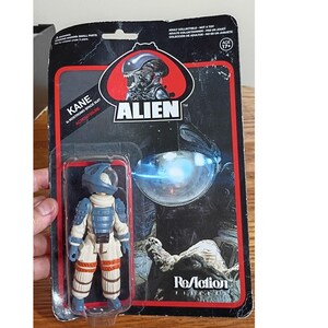 May include: A Kane in Nostromo space suit action figure from the Alien ReAction series, encased in original black and red blister packaging. The figure features a white suit with blue armor plating and orange leg bands, with the Alien logo visible.