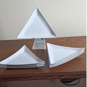 May include: A set of white triangular plates made of ceramic. One plate is displayed upright on a silver metal stand, while two stacks of additional triangular plates are positioned on either side of the stand, showcasing a uniform design.