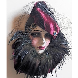May include: A decorative wall mask featuring a stylized face with dark makeup, adorned with a deep burgundy velvet headpiece, a black netting veil, a blue rhinestone accent, and a thick collar of dark, iridescent feathers surrounding the neck.