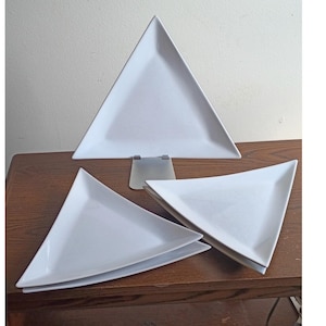 May include: A set of four white ceramic plates, each featuring a distinct triangular shape with smooth, flat surfaces and slightly raised edges. One plate is positioned upright on a metal stand, while the other three are stacked in two groups.
