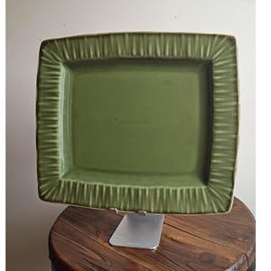May include: A rectangular ceramic serving platter in a solid sage green color. The platter features a smooth, flat center surface surrounded by a wide, raised border with a vertical ribbed or fluted texture along all four edges of the piece.