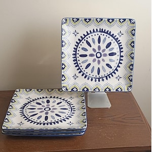 May include: A set of square ceramic plates featuring a central circular medallion pattern in shades of dark blue, light blue, and sage green. The border displays a repeating scalloped motif with small cross-shaped accents in matching colors.