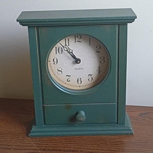 May include: A rectangular forest green wooden mantel clock with a flat top and base. The clock face is circular with a cream background, black Arabic numerals, and black hands. Below the clock face is a small drawer with a single round knob.