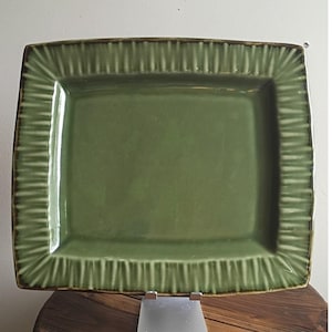 May include: A rectangular ceramic serving platter in a muted sage green glaze. The platter features a smooth, flat center basin surrounded by a wide, raised border decorated with a series of vertical, textured ridges along the entire perimeter.