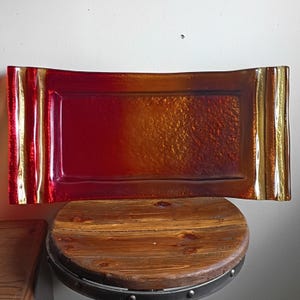 May include: A rectangular glass tray featuring a vibrant gradient of deep red and warm amber tones. The surface has a textured, pebbled finish in the center, while the two side edges are raised with a smooth, vertical ribbed pattern.