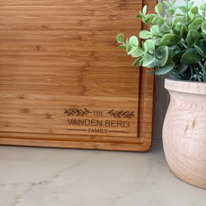 K&ouml;nnte beinhalten: Rechteckiges Bambus-Schneidebrett mit Saftrille und eingraviertem Text "THE VANDEN BERG FAMILY". Das hellbraune Brett liegt auf einer wei&szlig;en Marmoroberfl&auml;che. Eine kleine Topfpflanze befindet sich im Hintergrund.