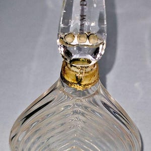 May include: A clear glass perfume bottle with a heart-shaped body featuring vertical ribbed detailing. The bottle has a gold-toned neck collar and a pointed, teardrop-shaped glass stopper with the text "AM DE" visible on its surface.