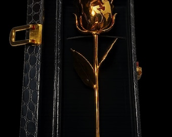 May include: A gold-plated rose in a black presentation box. The rose is fully detailed with petals, leaves, and stem, and is presented in a black box with a textured exterior and gold-colored clasp. The rose is a symbol of love and beauty.