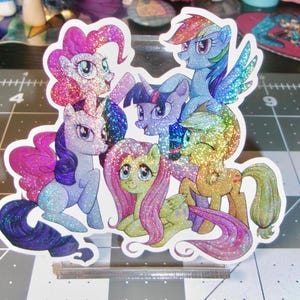May include: A die-cut holographic sticker featuring a group of six colorful cartoon ponies with large eyes and flowing manes. The characters are arranged in a cluster, displaying various poses and expressions, with a shimmering, glittery finish.