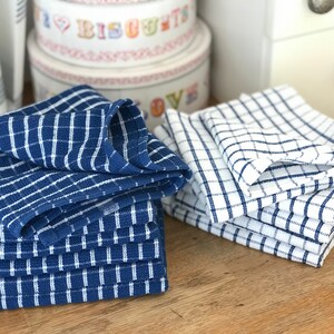 May include: Two stacks of blue and white checkered kitchen towels. The towels are folded and stacked on a wooden surface.
