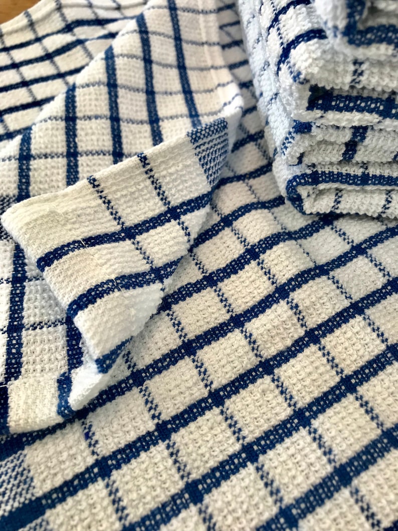 May include: A white kitchen towel with a blue checkered pattern. The towel is folded and stacked on top of another towel.