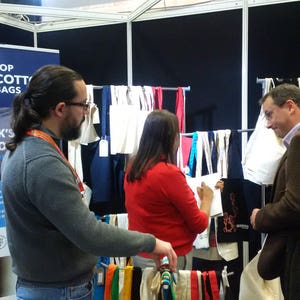 May include: A trade show booth with a sign that reads "Top Cotton Bags, The UK's Cotton Bag Manufacturer". There are several white cotton tote bags hanging on a rack, along with colorful fabric samples. Two people are standing at the booth, looking at the bags.