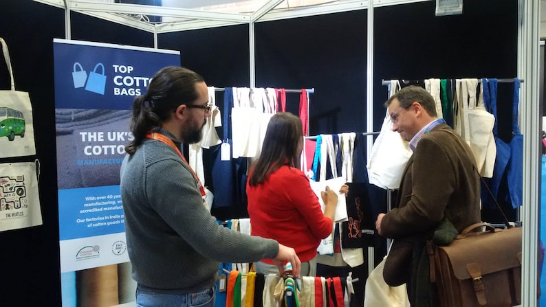 May include: A trade show booth with a sign that reads "Top Cotton Bags, The UK's Cotton Bag Manufacturer". There are several white cotton tote bags hanging on a rack, along with colorful fabric samples. Two people are standing at the booth, looking at the bags.