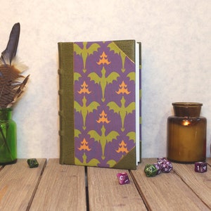 May include: A purple and green book with a dragon pattern on the cover, a green glass jar with feathers, a small picture frame with a butterfly, and a few dice on a wooden table.
