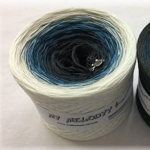 Puede incluir: Una gran madeja de hilo multicolor con un degradado de blanco, azul y negro. El hilo est&aacute; etiquetado como "MY MELODYY" y tiene un dije de nota musical. La direcci&oacute;n del sitio web www.wolltraum.ch/com tambi&eacute;n es visible. Ideal para proyectos de tejido y ganchillo.