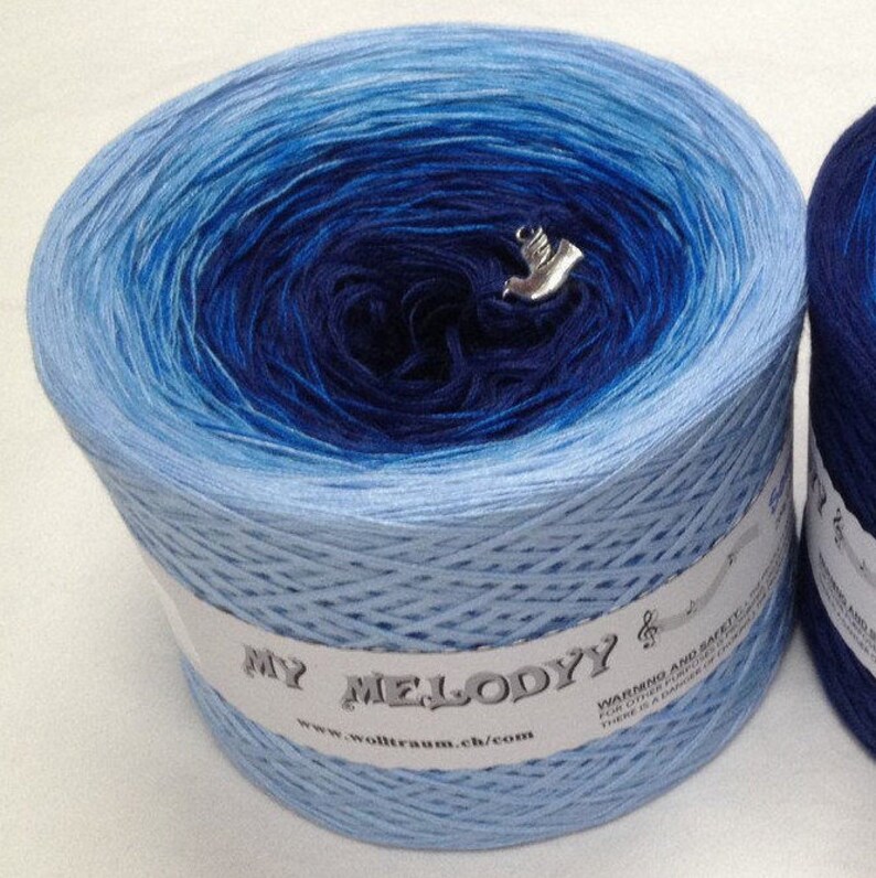 K&ouml;nnte beinhalten: Eine Kn&auml;uel blaues Ombre-Garn mit einem wei&szlig;en Etikett, auf dem "My Melody" und eine Website-Adresse stehen. Das Garn ist zu einer Kugel gewickelt und hat einen silbernen Anh&auml;nger.