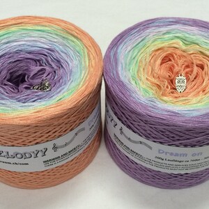 May include: Two skeins of colorful yarn in a gradient of colors, including peach, purple, green, and yellow. Each skein has a small charm and a label with text. The yarn is likely for knitting or crochet projects.