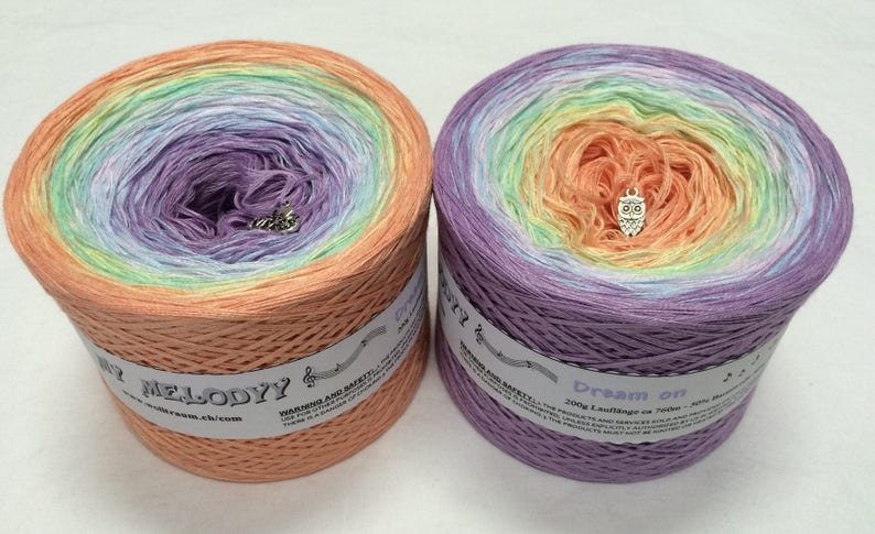 May include: Two skeins of colorful yarn in a gradient of colors, including peach, purple, green, and yellow. Each skein has a small charm and a label with text. The yarn is likely for knitting or crochet projects.
