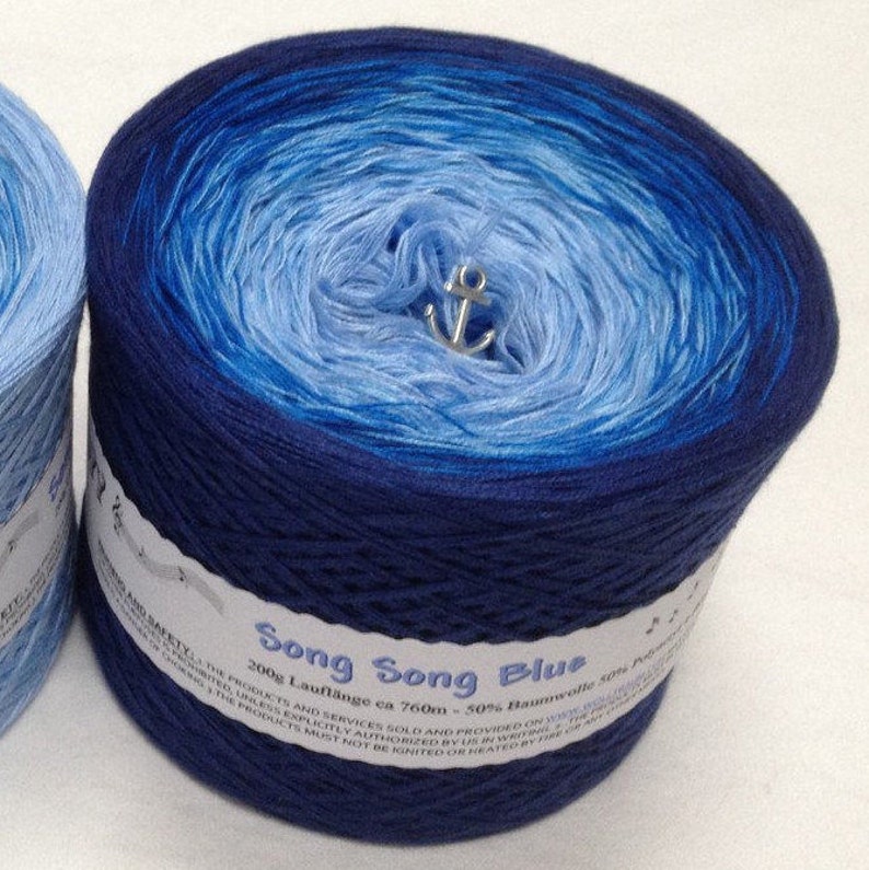 K&ouml;nnte beinhalten: Eine Kn&auml;uel blaues Ombre-Garn namens "Song Song Blue" mit einem wei&szlig;en Etikett, auf dem "200g Laufl&auml;nge ca 760m - 50% Baumwolle 50% Polyacryl" steht.