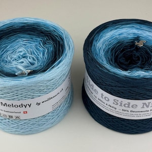 May include: Two cylindrical cakes of gradient yarn. The left cake transitions from dark navy to light blue. The right cake transitions from light blue to dark navy. Each features a white paper label with text including My Melodyy and Side to Side Nr. 24.