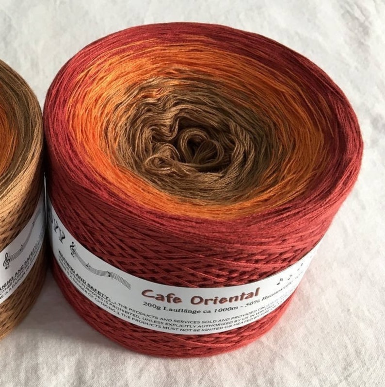 May include: A skein of yarn in a gradient of red, orange, and brown colors. The yarn is labeled "Cafe Oriental" and "200g Lauflange ca 1000m - 50% Baumwolle".