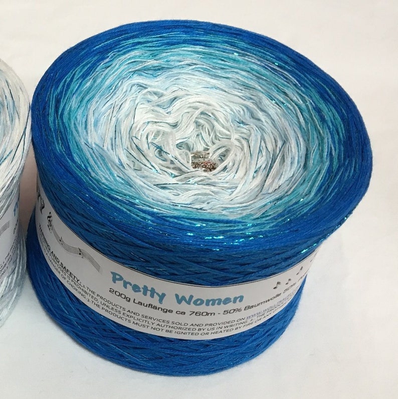 May include: A skein of blue, teal, and white yarn with a glittery sheen. The yarn is labeled "Pretty Women 200g Lauflange ca 760m - 50% Baumwolle"
