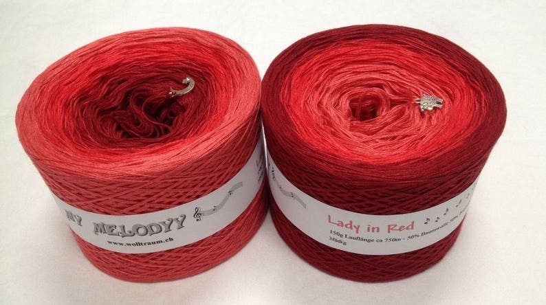 May include: Two skeins of red yarn, one labelled "Melody" and the other "Lady in Red". Both skeins are ombre, transitioning from a lighter red to a darker red.