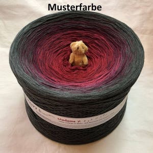 May include: A skein of yarn in a gradient colorway, starting with a dark gray and transitioning to a deep red. The yarn is wound into a ball shape and a small teddy bear is sitting on top of the yarn. The label on the yarn reads "Madame X 500g Lauflänge ca 3800m 50% Baumwolle 50% Polyacryl"