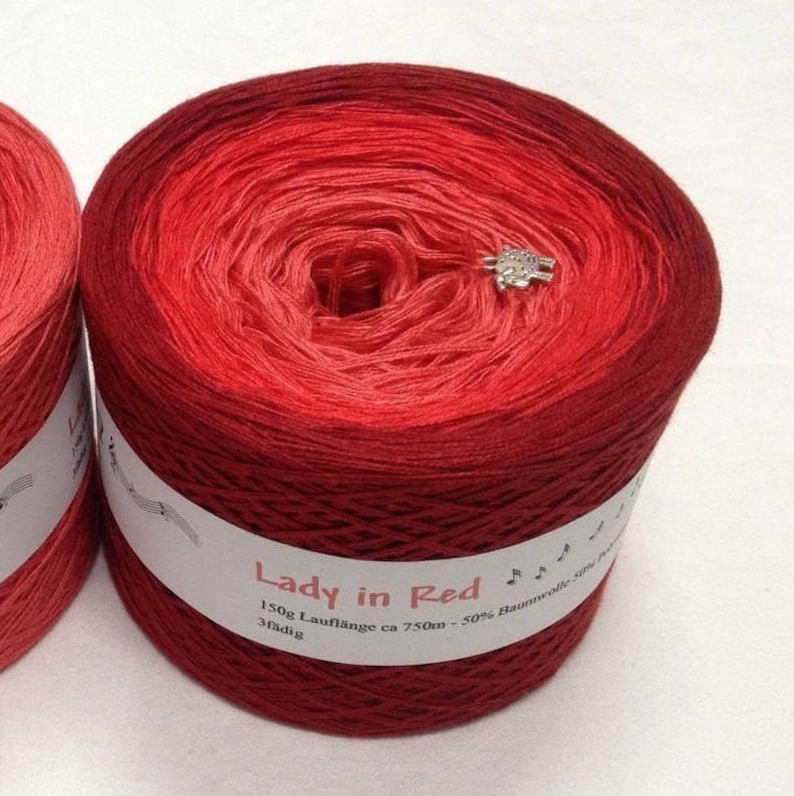 May include: A skein of red yarn with a gradient from a lighter red to a darker red. The yarn is labelled "Lady in Red" and has a label with the text "150g Laufl&auml;nge ca 750m - 50% Baumwolle 3f&auml;dig".