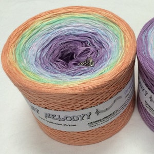 May include: A large, colorful yarn cake with a gradient of peach, yellow, green, blue, and purple. The yarn is labeled "My Melodyy" and includes a small charm. The yarn is for knitting and crochet projects.