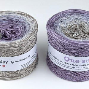 May include: Two cylindrical yarn skeins. One is labeled "My Melodyy" and the other "Que sera". The yarn is a gradient of gray, purple, and white. Each skein has a white label with text and a small charm.