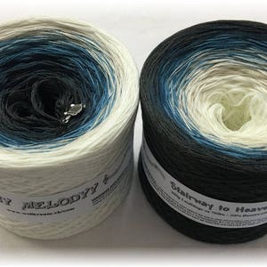 Puede incluir: Dos madejas de hilo con un esquema de color degradado. Una es blanca con azul y negro, la otra es negra con azul y blanco. Cada una tiene una etiqueta y un sujetahilos plateado.